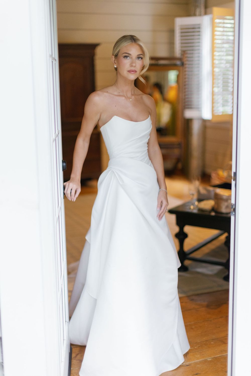 Bride in an elegant strapless white satin wedding gown with draped skirt stands in a doorway of a bright, rustic wooden room.