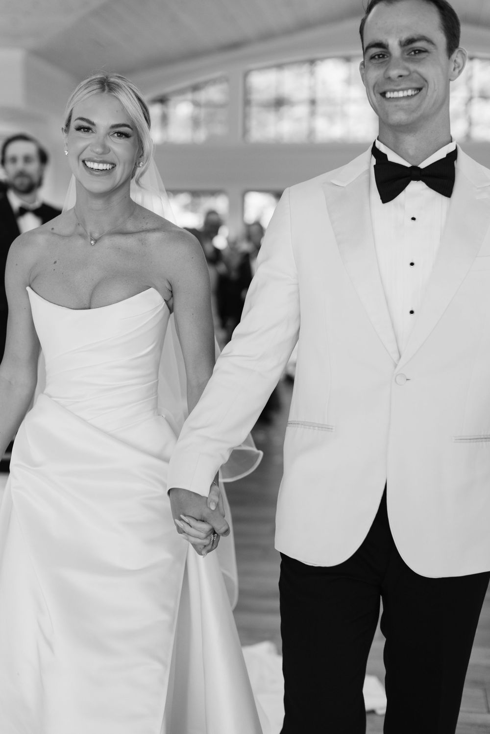 Smiling bride in a strapless satin gown and veil holds hands with a groom in a white dinner jacket and bow tie in a black and white photo.