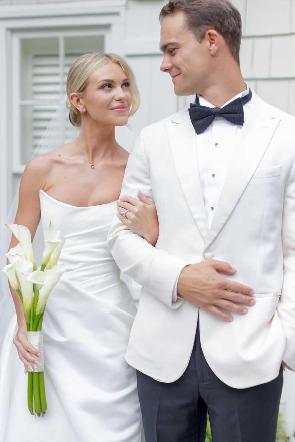 Smiling bride in a strapless white gown holding calla lilies stands arm in arm with a groom in a white dinner jacket and black bow tie.