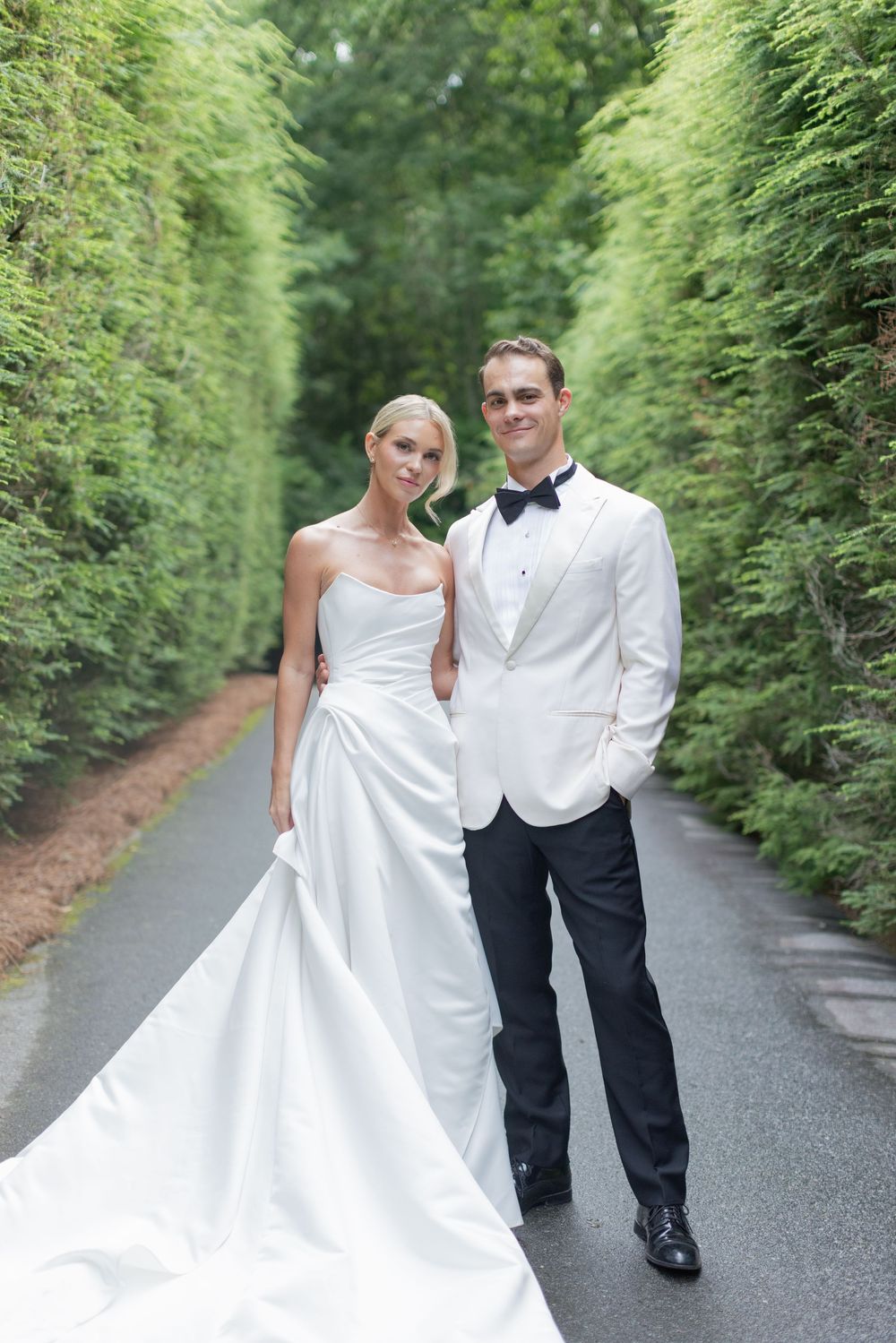 Bride in a strapless white satin gown and groom in a white dinner jacket and bow tie pose on a path between tall green hedges.