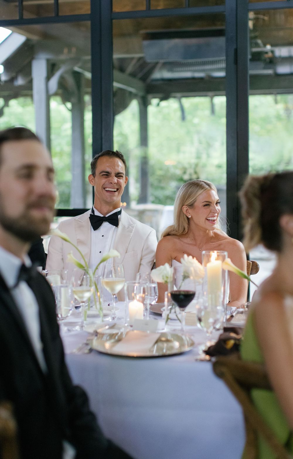 Smiling couple in formal attire seated at a candlelit wedding reception table with glassware and flowers in a bright glass-walled venue.