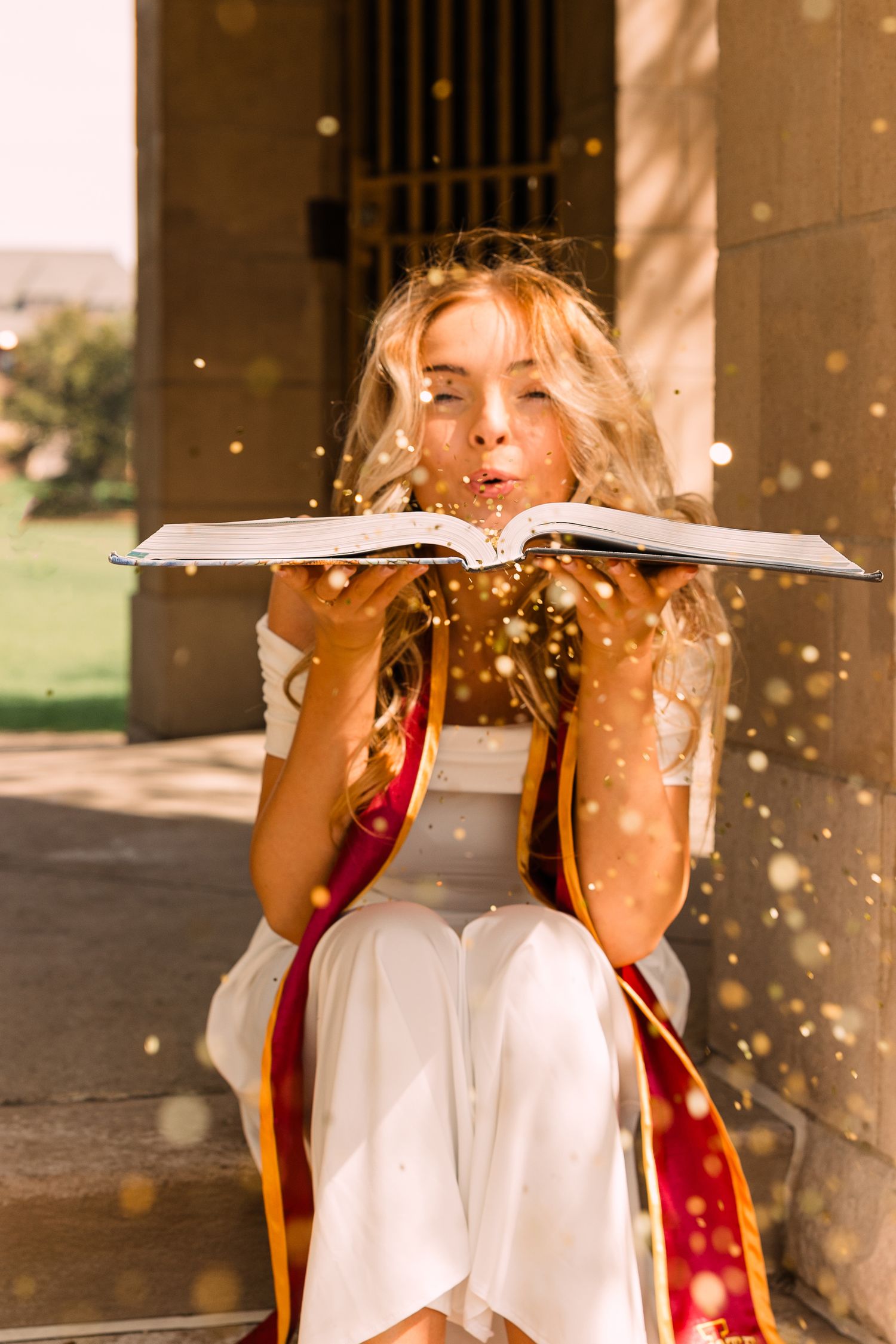 Graduate in a white dress and maroon and gold sash blows glitter from an open book while sitting in a sunlit archway.