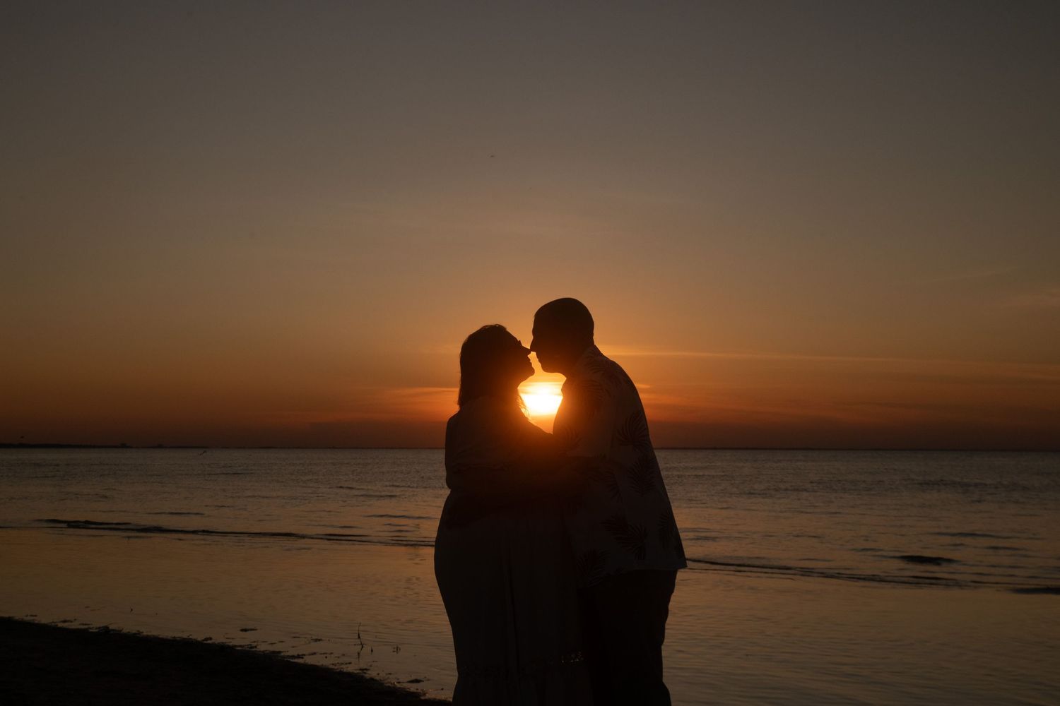Silhouette of a romantic couple standing together on a beach at sunset with glowing orange sky.