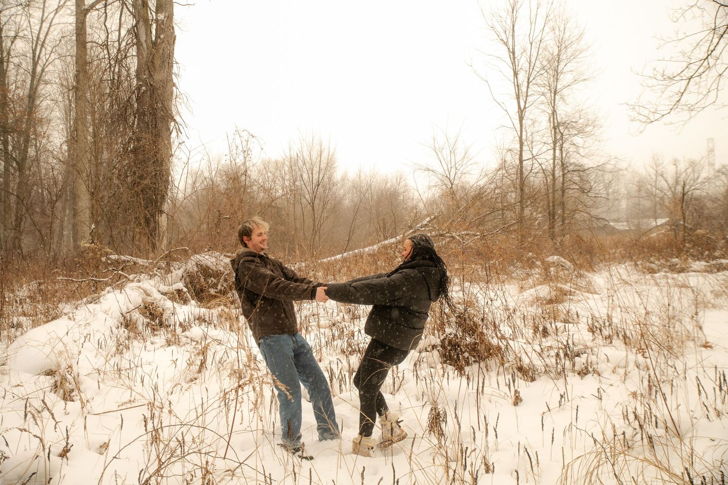 Couple dancing playfully in a snowy winter forest clearing, wearing casual winter clothes among bare trees.