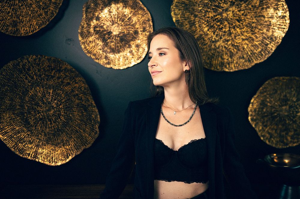 Woman in black blazer and lace top poses against dark wall with gold decorative disc accents.