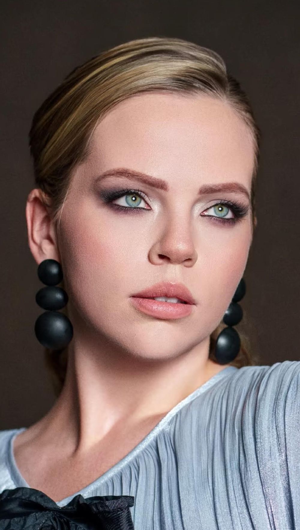 Young woman with green eyes, smoky eye makeup, slicked-back blonde hair, and black spherical drop earrings wearing a light blue top.
