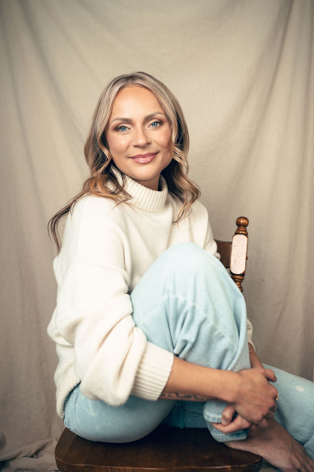 Smiling woman in white turtleneck sweater and light blue jeans sitting relaxed on wooden chair against neutral backdrop.