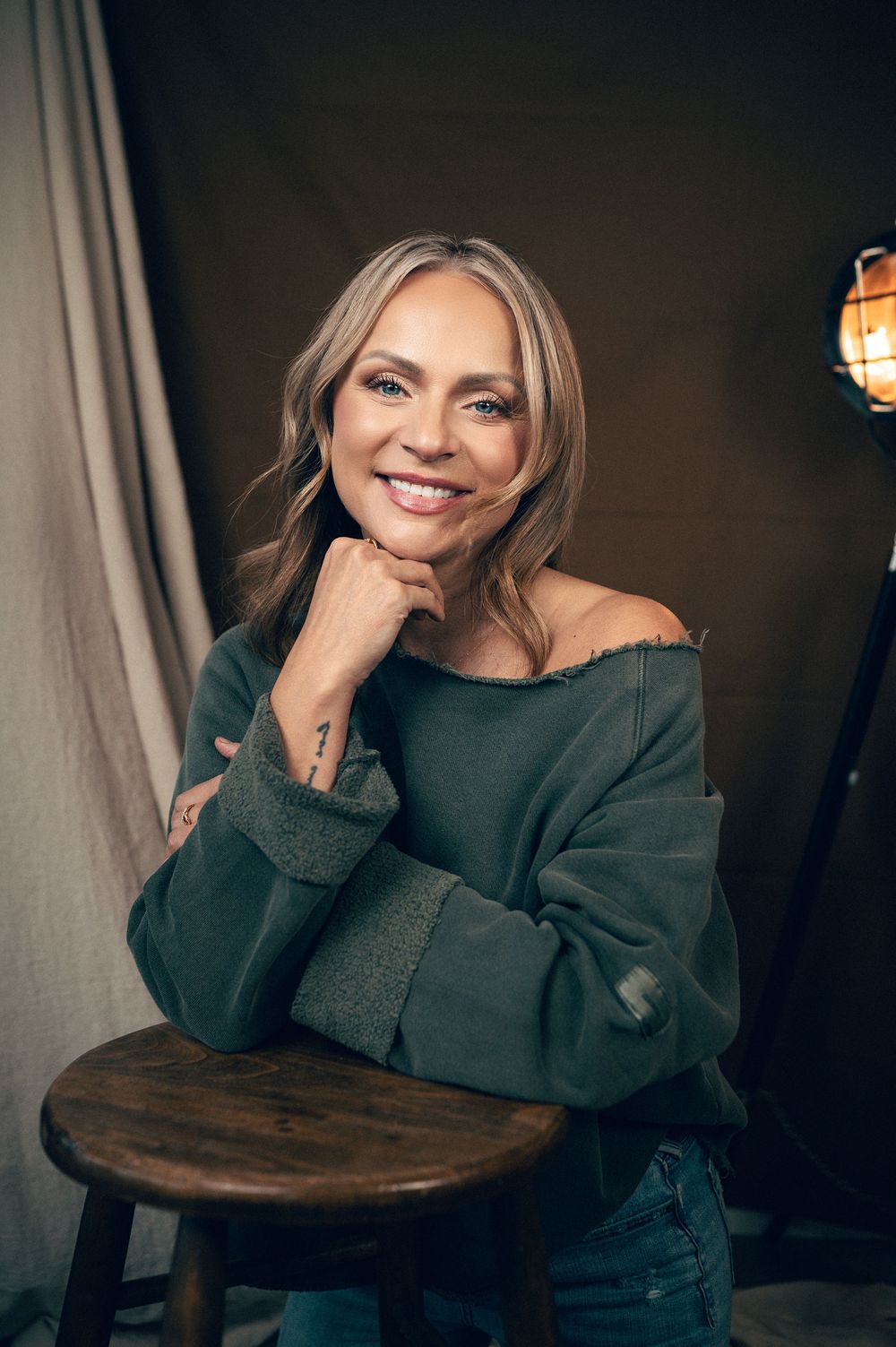 Smiling woman with blonde hair wearing an off-shoulder green sweatshirt, leaning on a wooden stool in a studio setting.