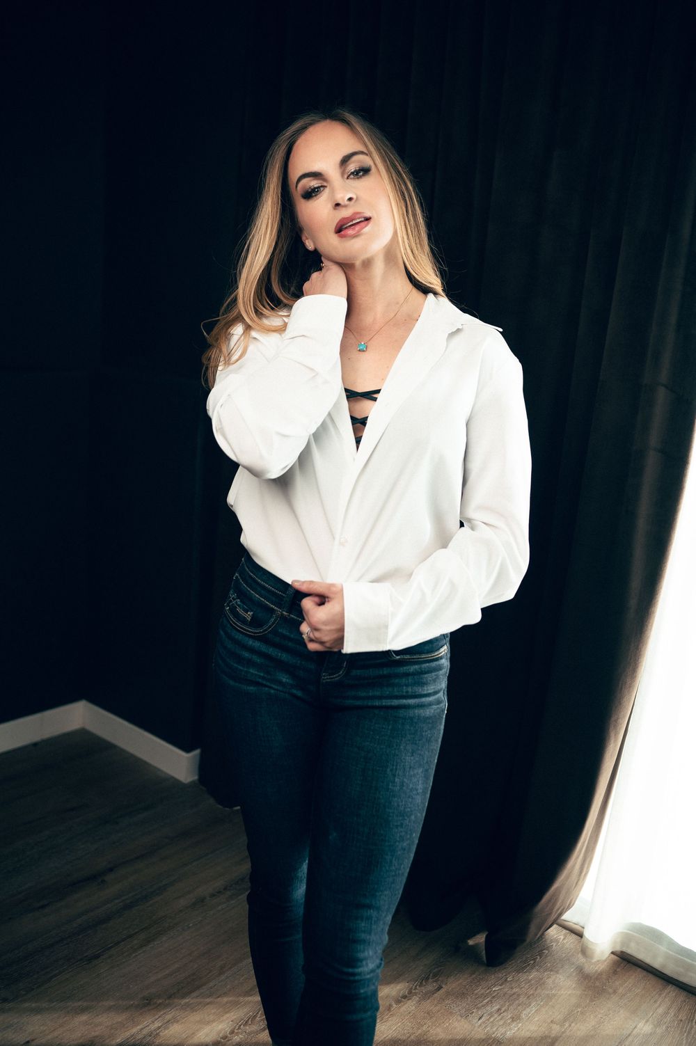 A blonde woman poses confidently in a white button-up blouse and dark skinny jeans against a black curtain backdrop.