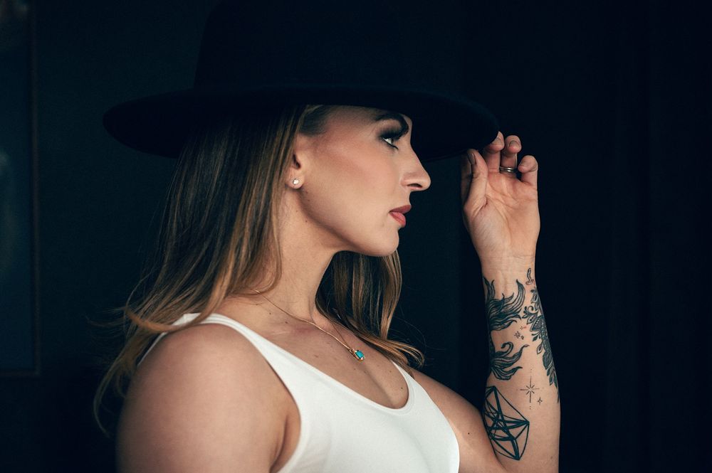 Woman in white top and black wide-brim hat with floral arm tattoo posing in profile against dark background.