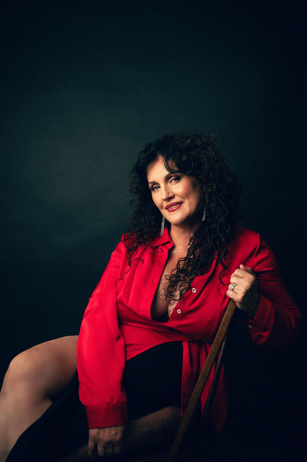 A confident woman with curly dark hair poses in a red shirt against a dark background, holding a wooden prop.