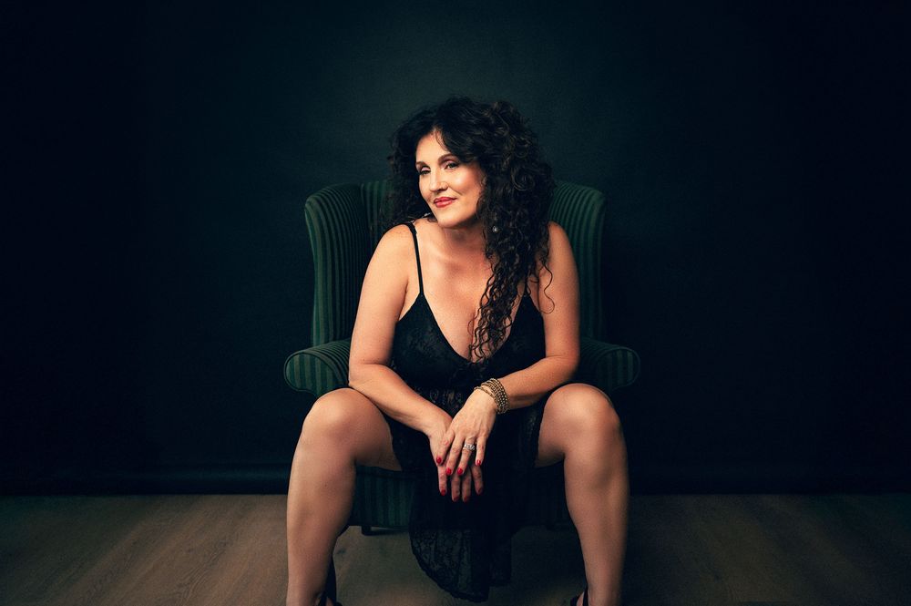 Woman with curly hair in black dress sits elegantly on green velvet chair against dark studio background.
