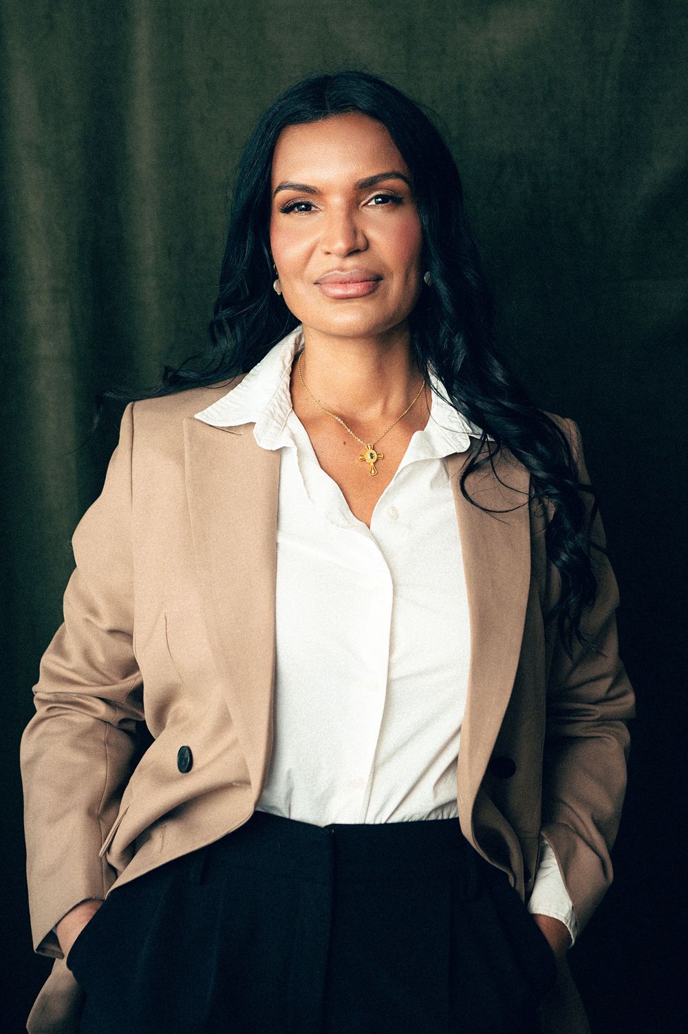 Professional woman in beige blazer and white shirt poses confidently against dark background wearing gold necklace.