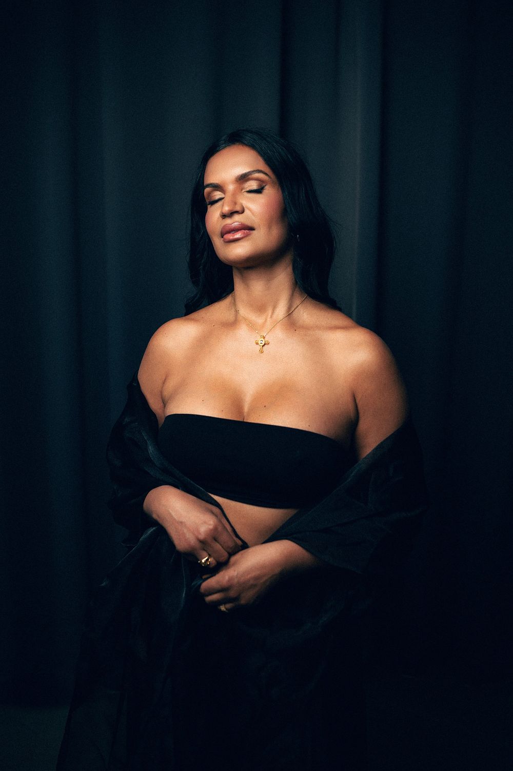 Woman in black strapless top and gold necklace poses elegantly against dark curtain backdrop.