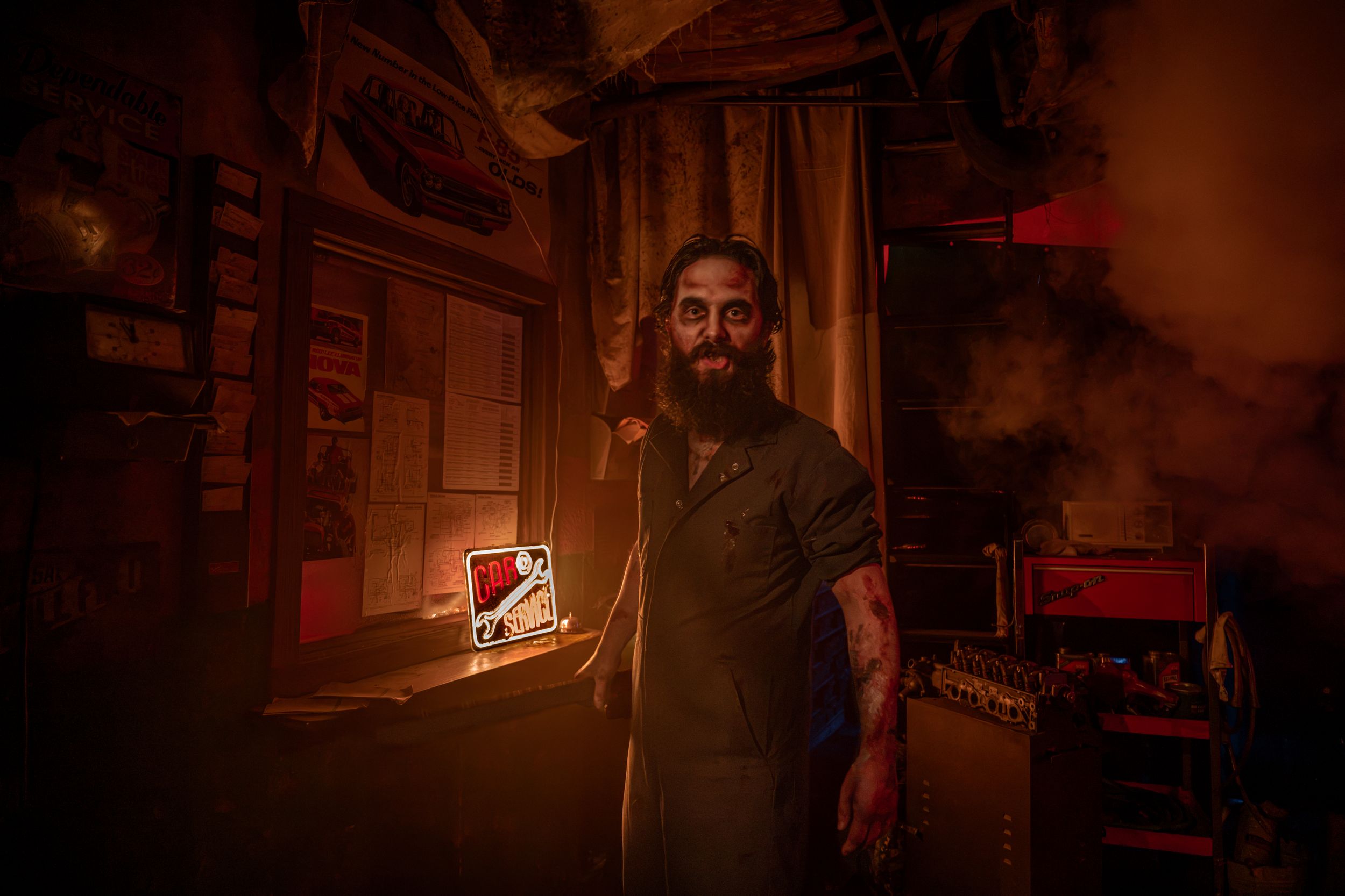 Bearded man in dark apron stands in smoky, dimly lit workshop with red-orange atmospheric lighting.