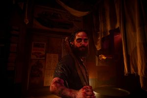 Bearded tattooed man sitting in dimly lit room with red and amber lighting, looking intensely to the side.