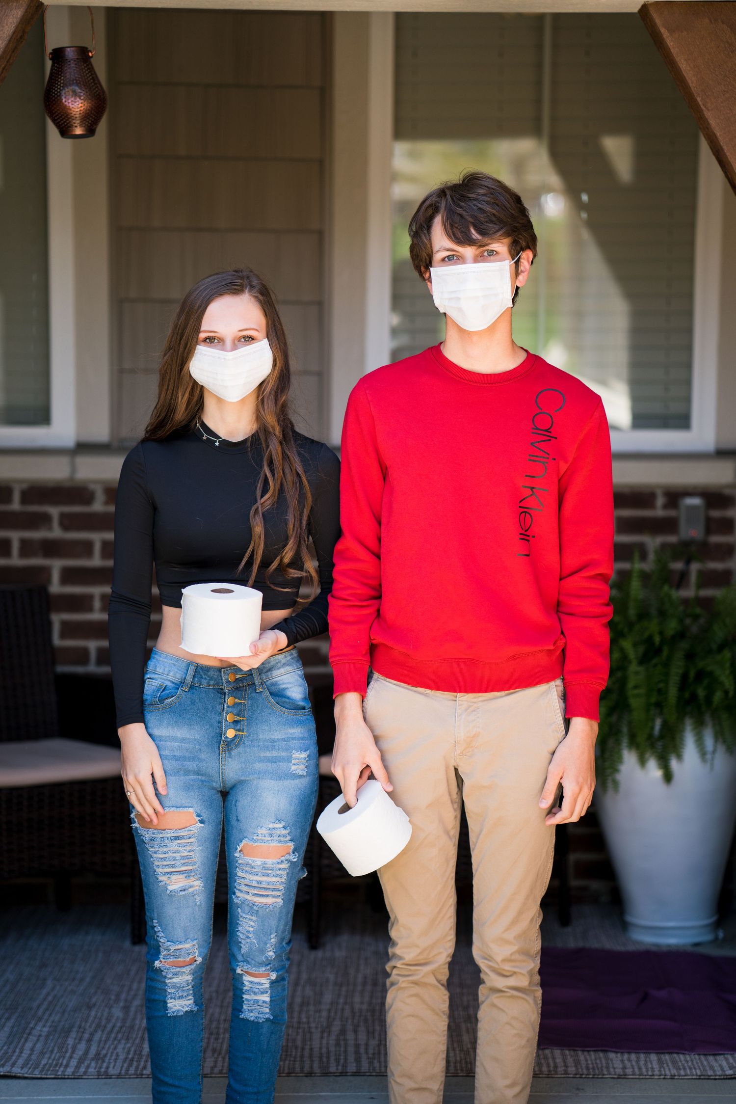 Two young people wearing face masks holding toilet paper rolls while standing on a house porch during COVID-19.
