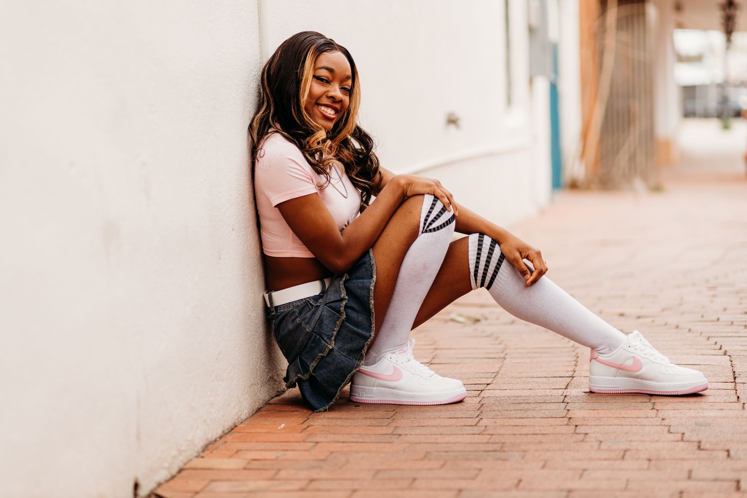 Smiling young woman in a pink crop top, denim skirt, striped knee-high socks and white sneakers sits against a wall on a brick sidewalk.