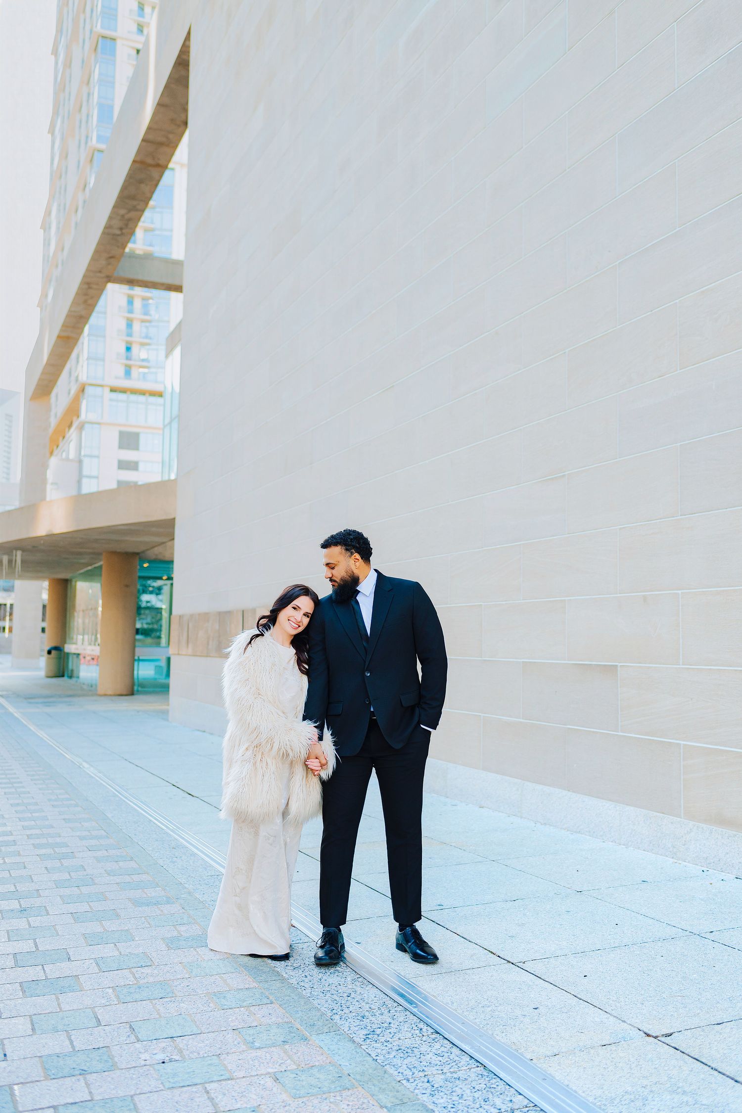 Couple in formal attire poses outside modern building, woman in white fur coat, man in black suit holding hands.