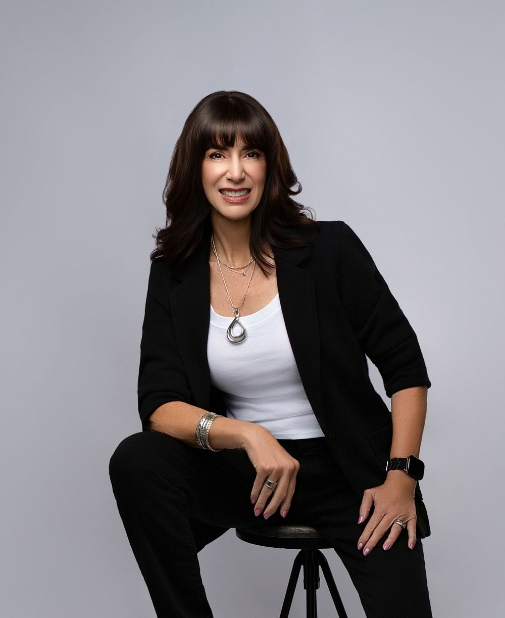 Woman with dark hair in a black blazer seated on a gray backdrop, corporate headshot for women
