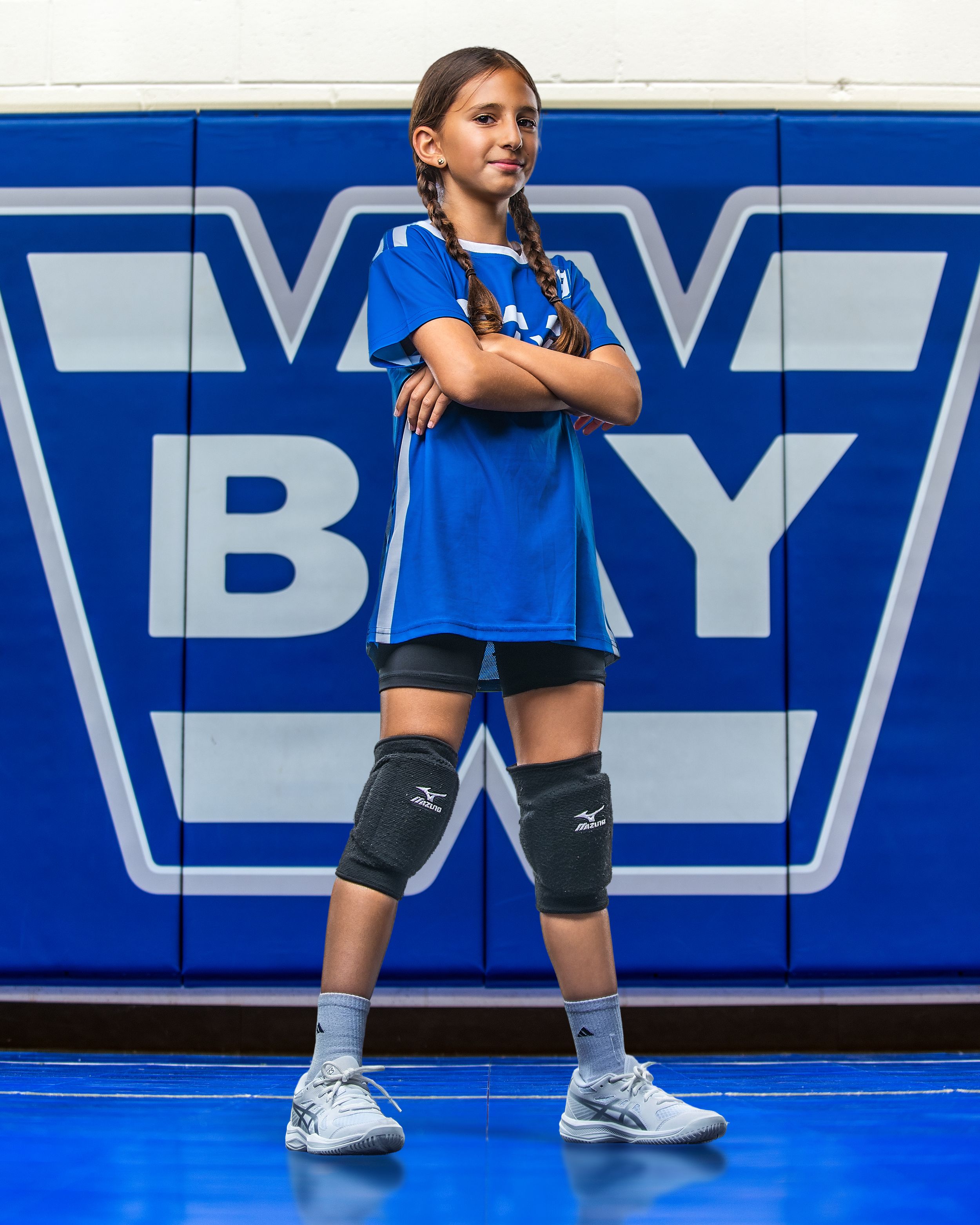 Young girl in blue volleyball uniform with knee pads stands confidently arms crossed in front of a blue gym wall.