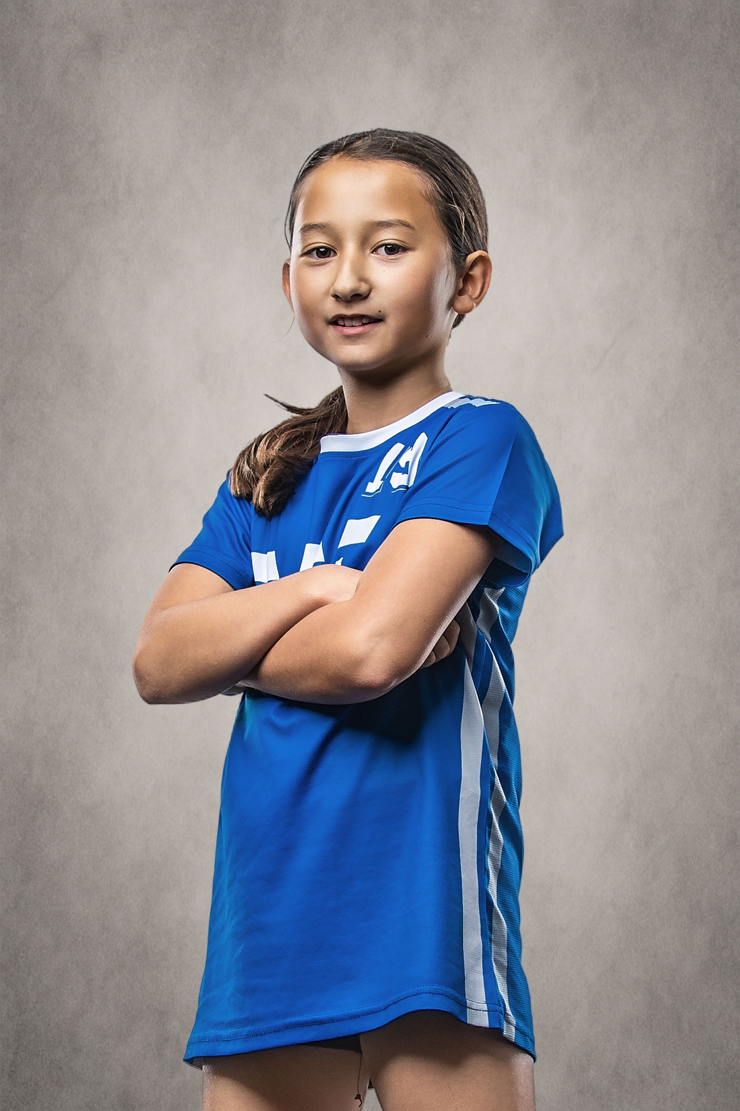 Young girl in blue soccer jersey poses confidently with arms crossed for a sports team portrait photo.
