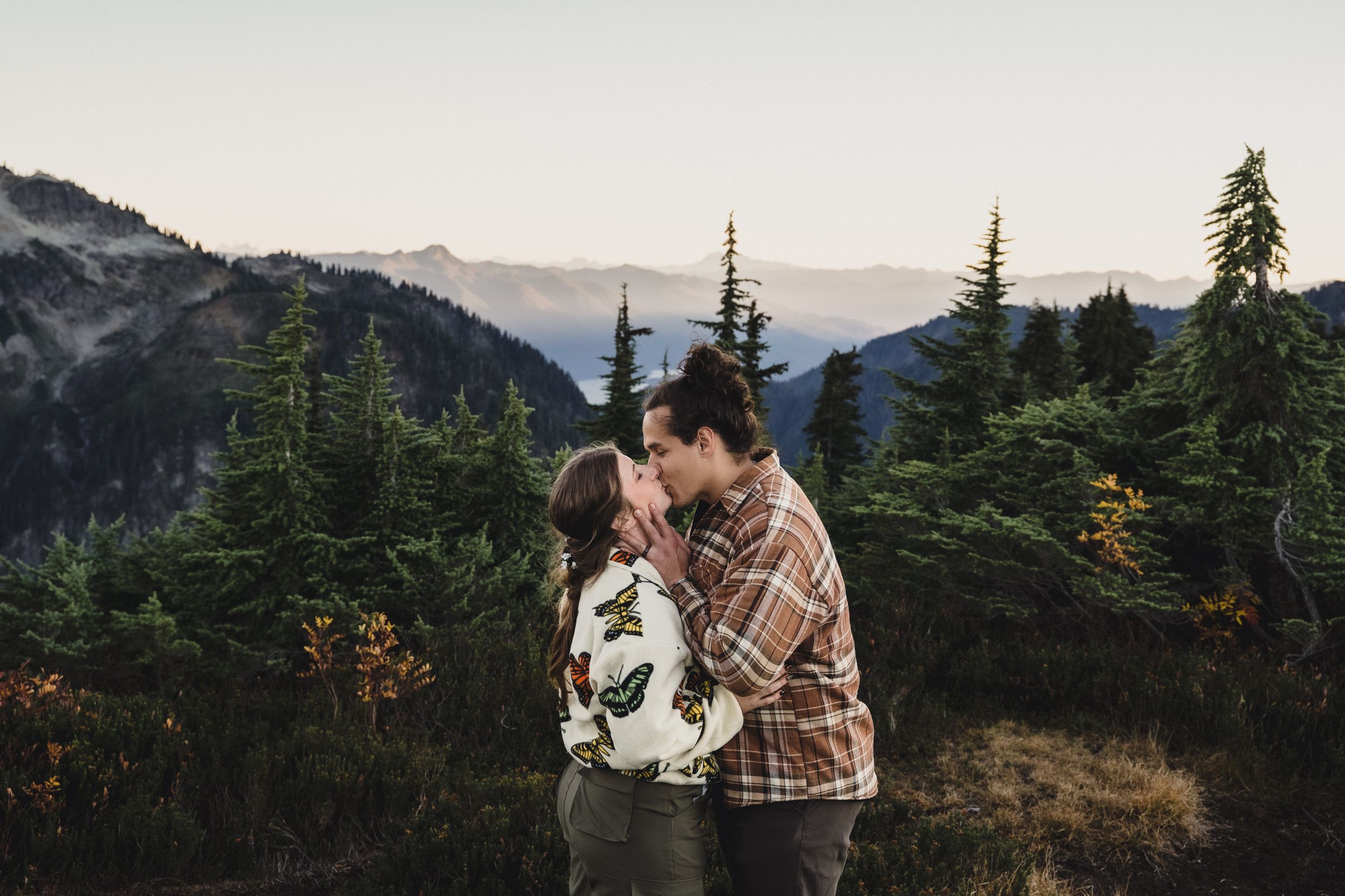 Couple in plaid and floral jackets sharing a kiss on a mountain meadow with evergreen trees and rugged peaks in the background.