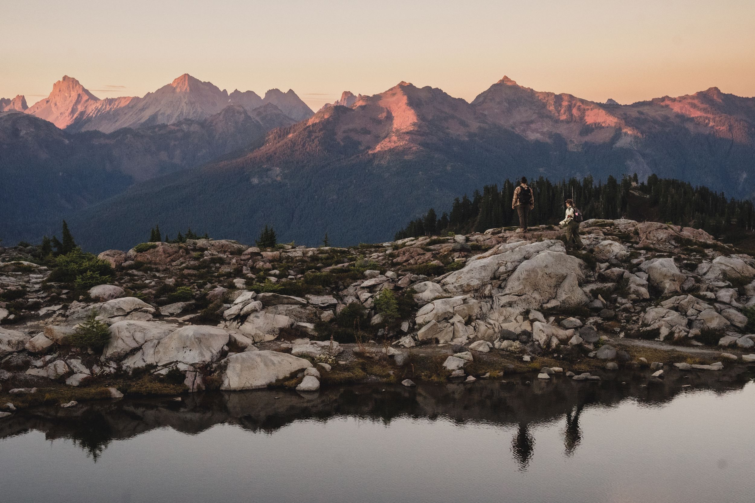 Two hikers stand on rocky alpine terrain beside a still mountain lake reflecting pink sunset light on jagged peaks.
