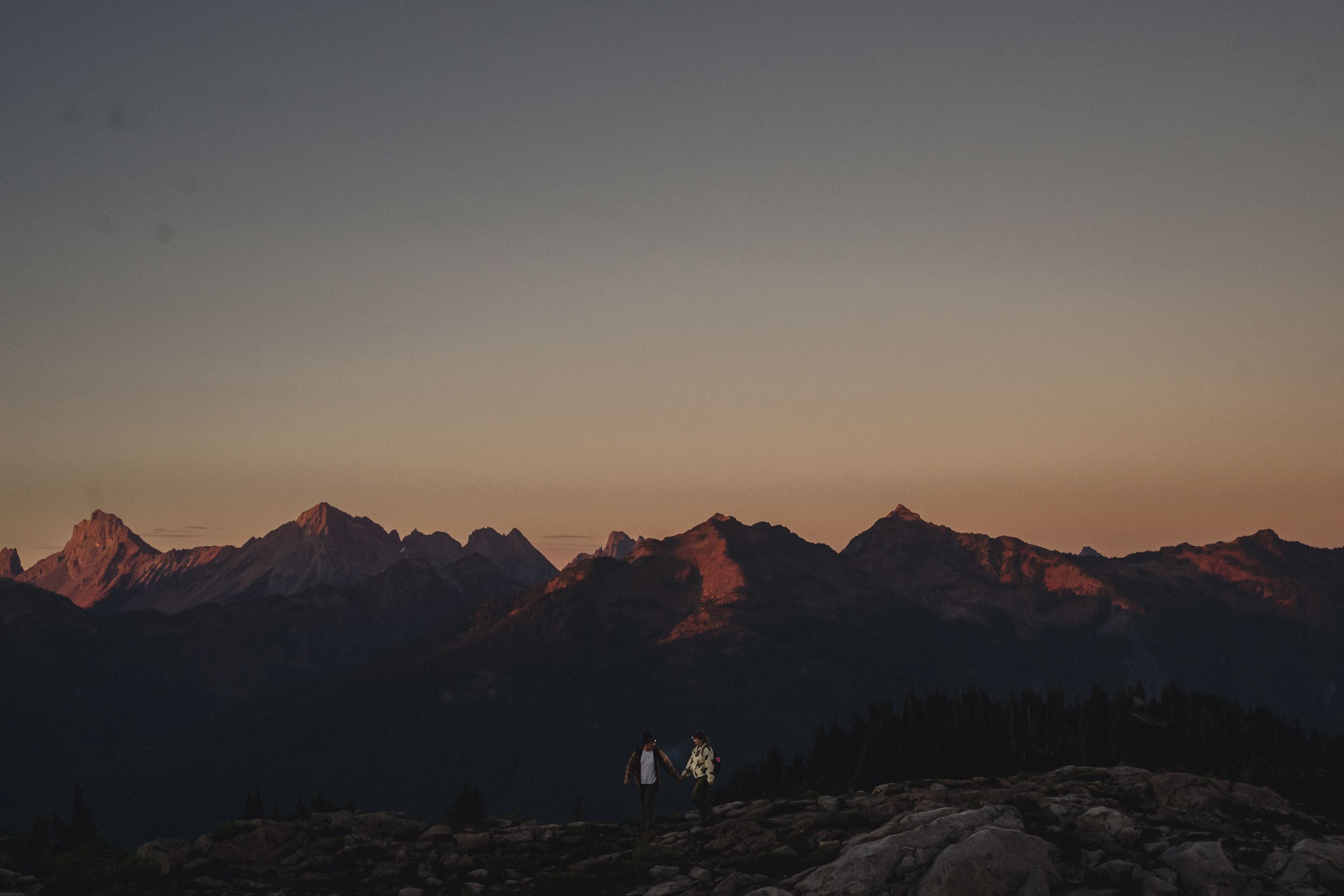 Mountain range glowing with pink alpenglow at dusk, with a small cabin and two people on a rocky ridge in the foreground.