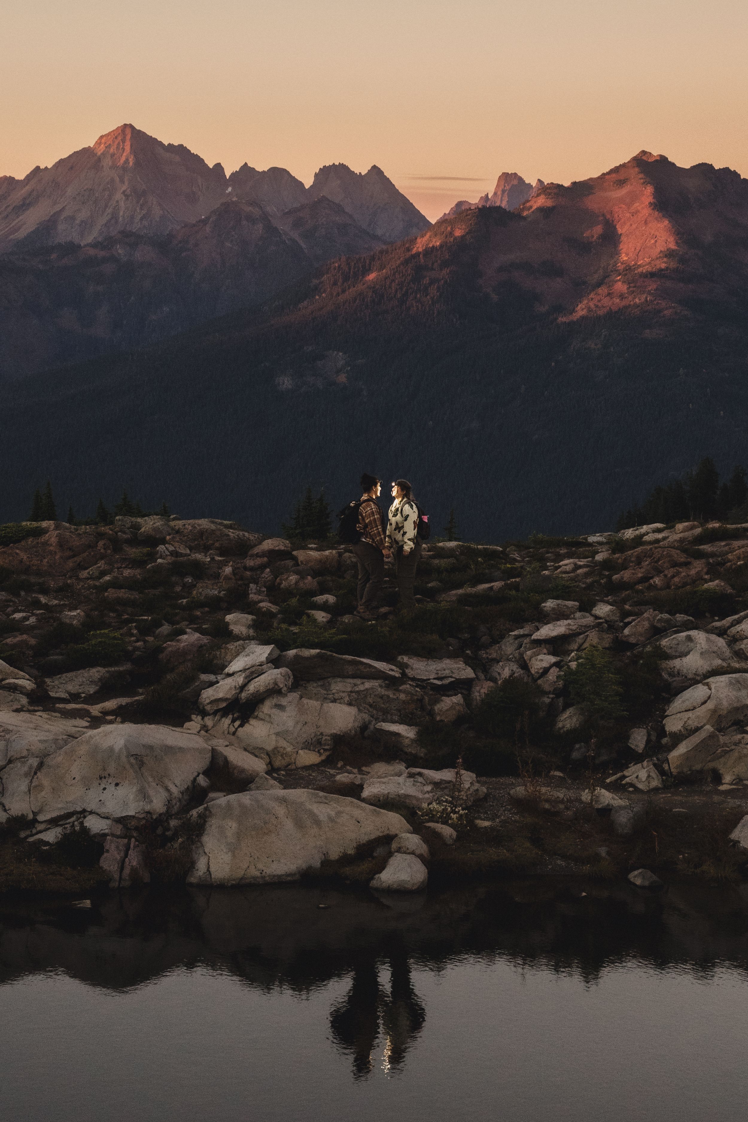 Two hikers stand on rocky alpine terrain beside a still mountain lake at sunset, with their reflection and glowing peaks behind them.