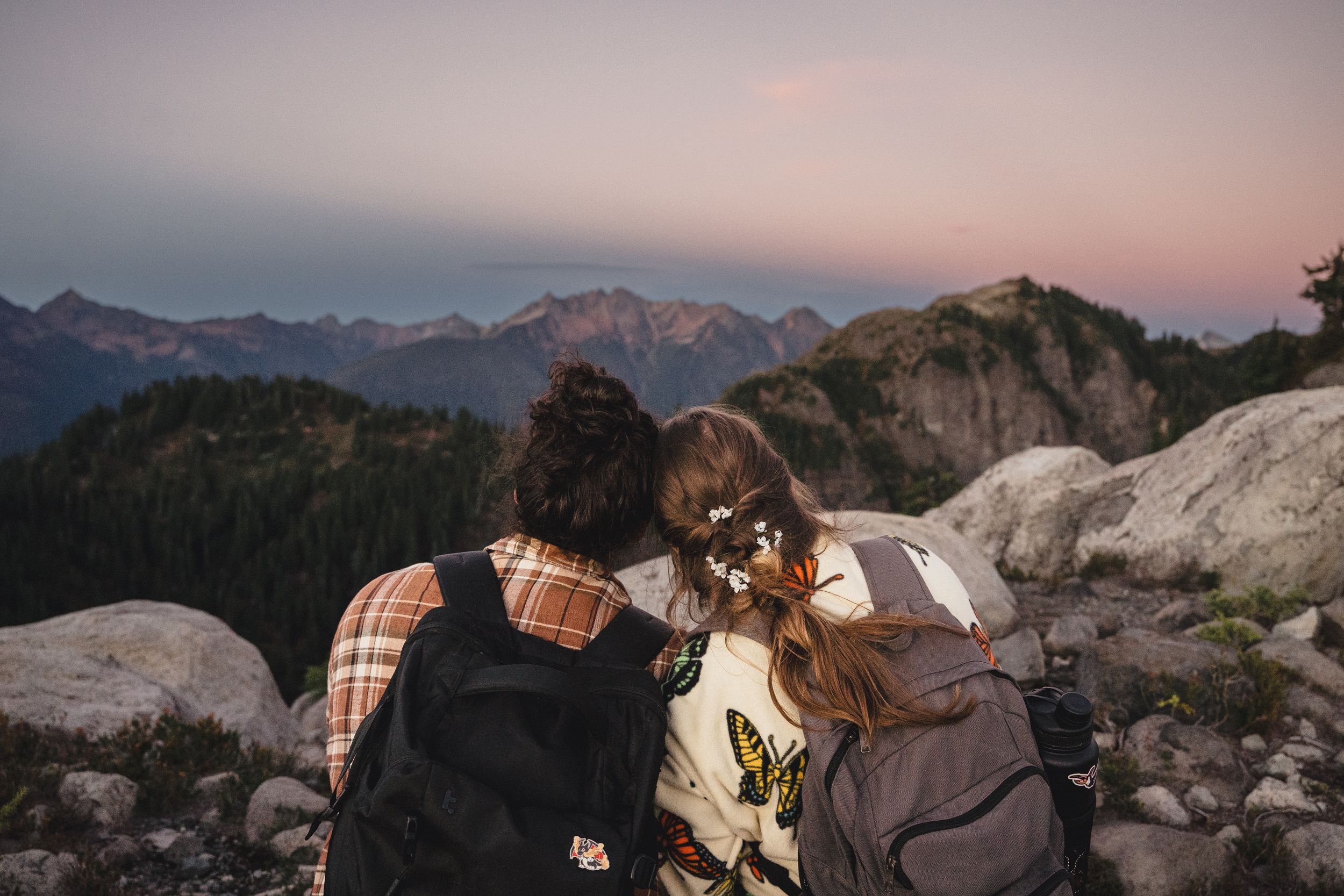 Two hikers with backpacks sit on rocky terrain, gazing at a pink and purple sunset over rugged mountain peaks and forested valleys.