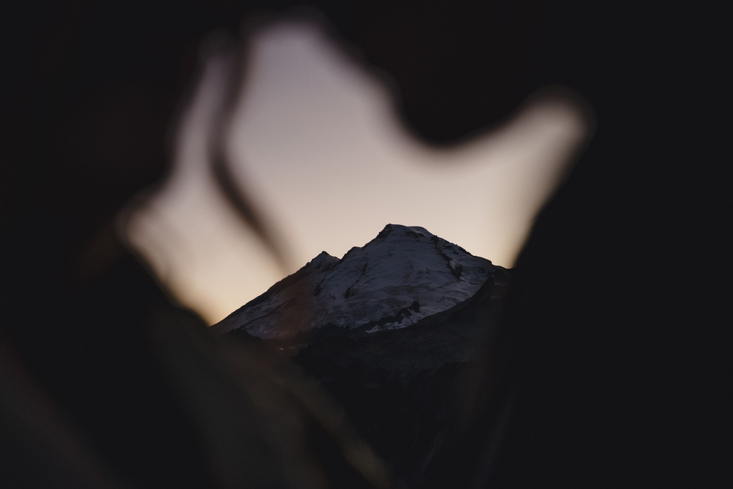 Snow-capped mountain peak at dusk framed through a dark blurred silhouette opening, with a soft glowing twilight sky behind.