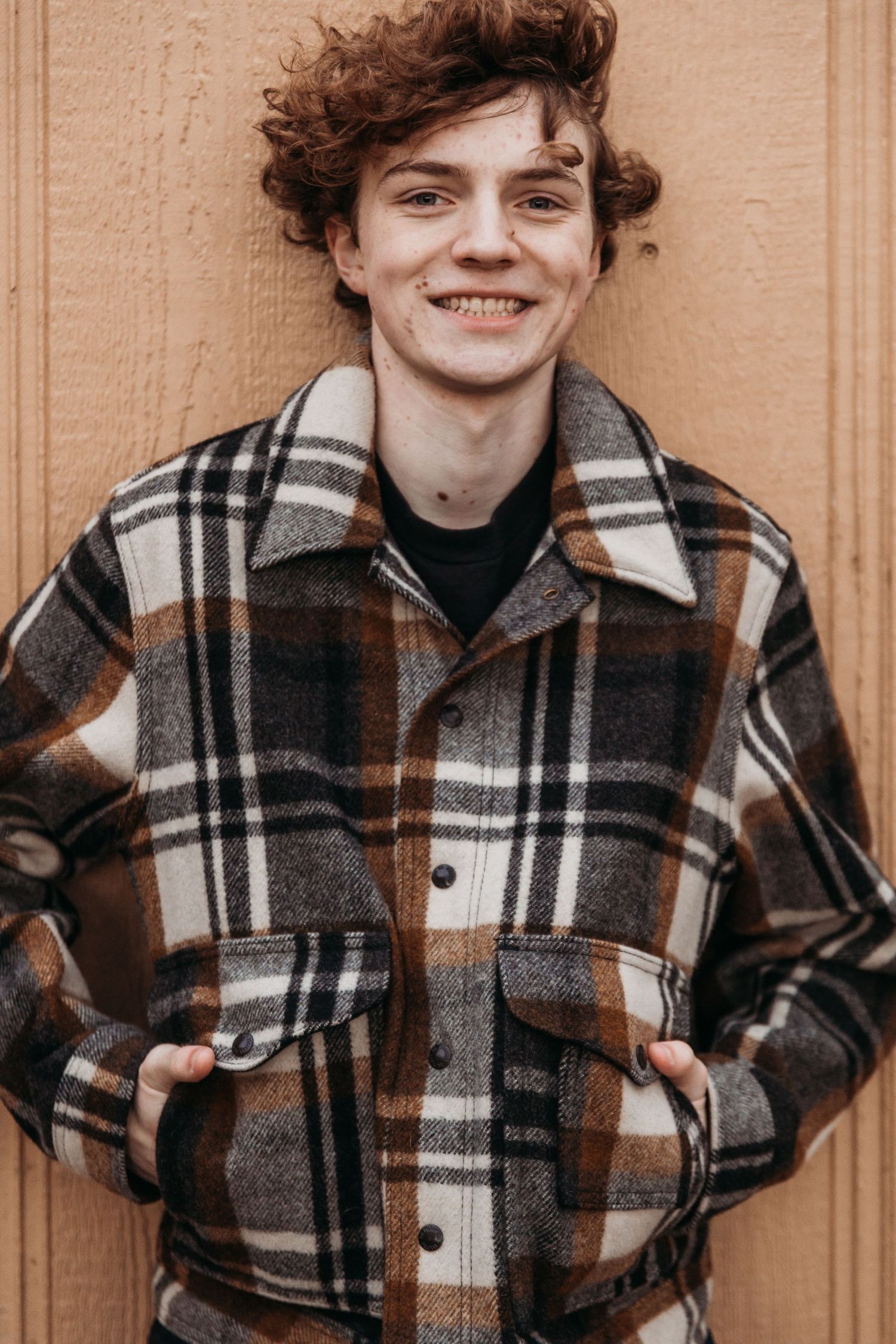 Smiling young man with curly auburn hair wearing a brown and cream plaid flannel jacket, standing against a light wood panel wall.