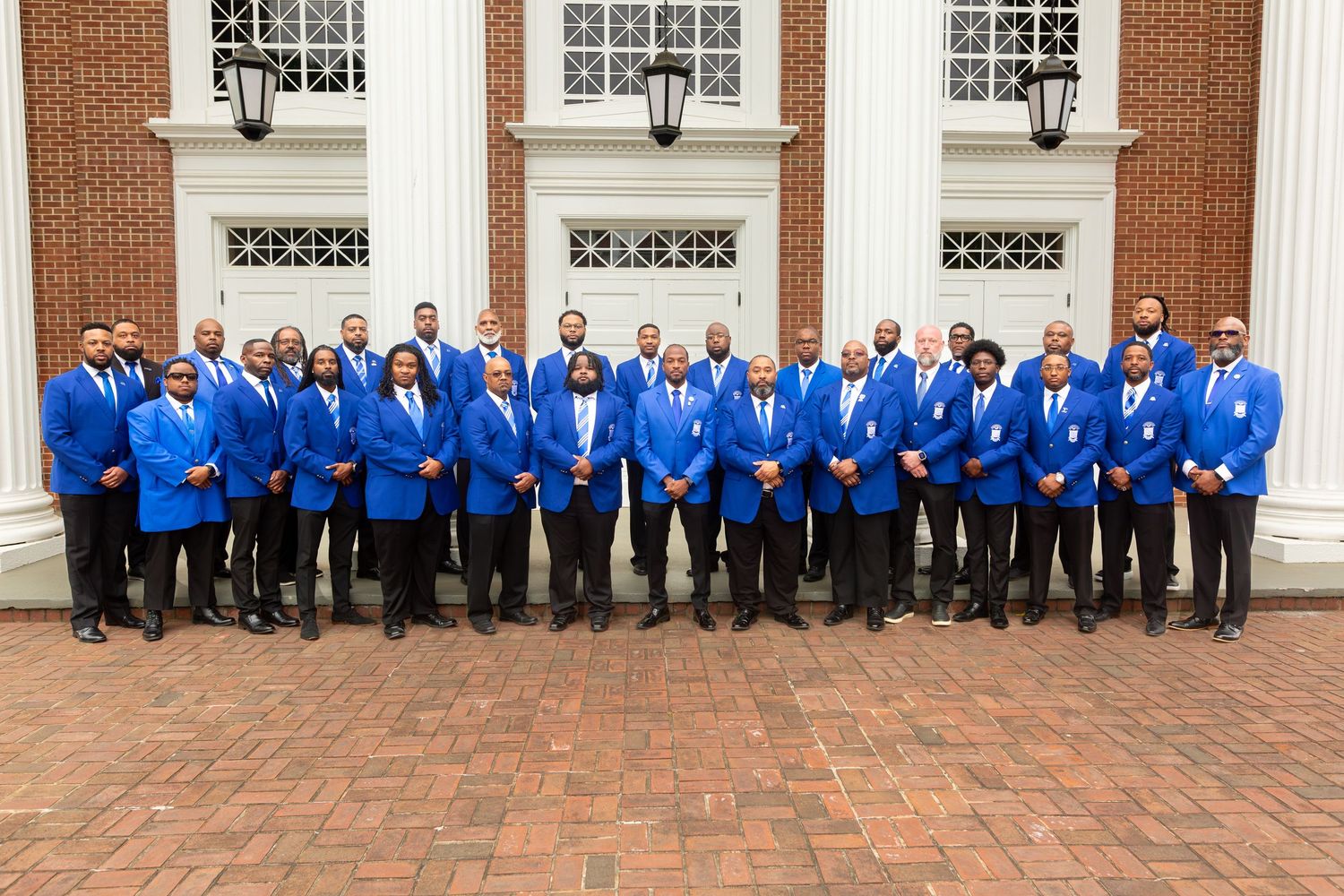 Group of men in matching royal blue blazers and dark trousers pose in rows on a brick patio in front of a brick building with white columns.