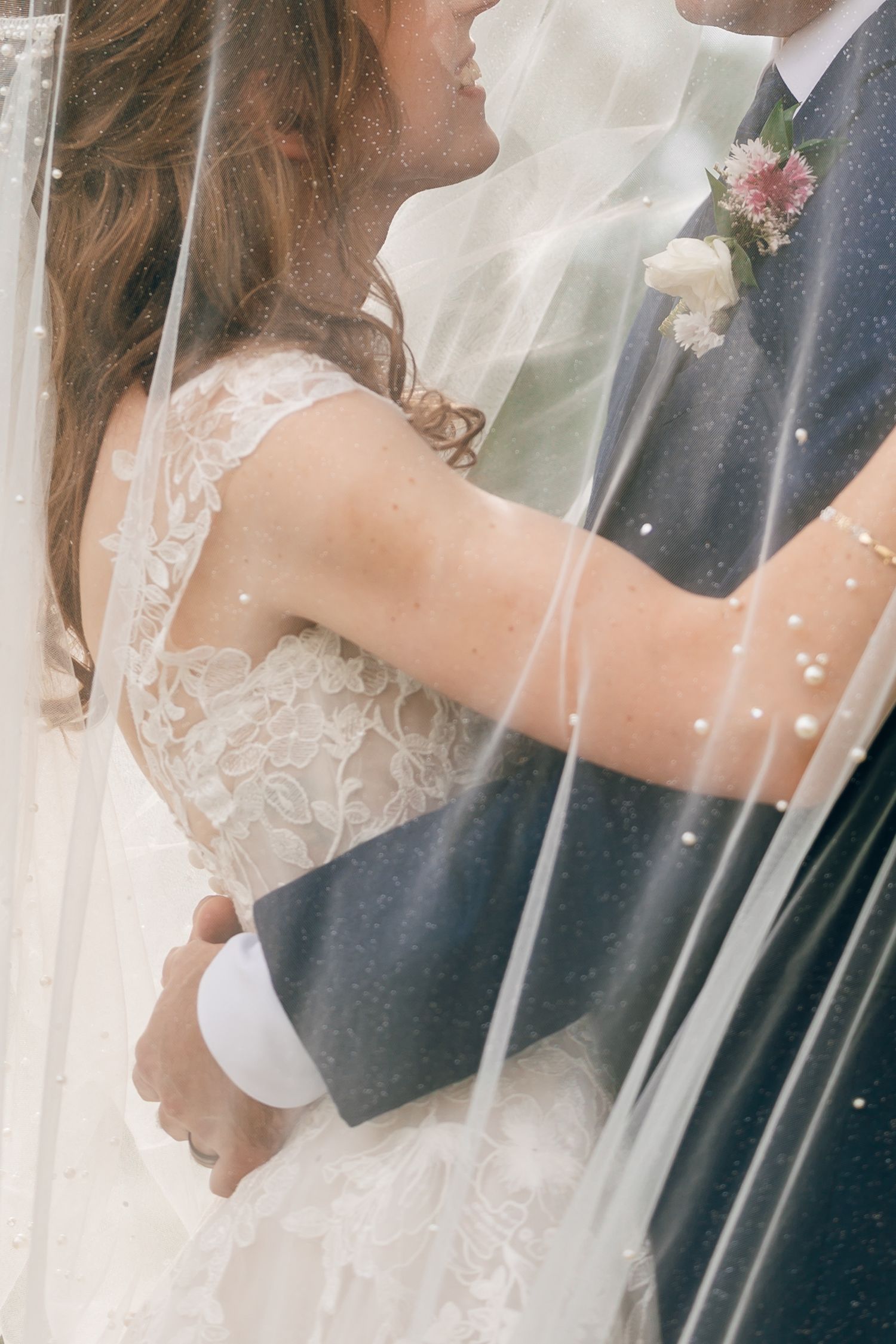 Bride in a lace wedding gown embracing a groom in a navy suit with a floral boutonniere, framed by a sheer beaded veil.
