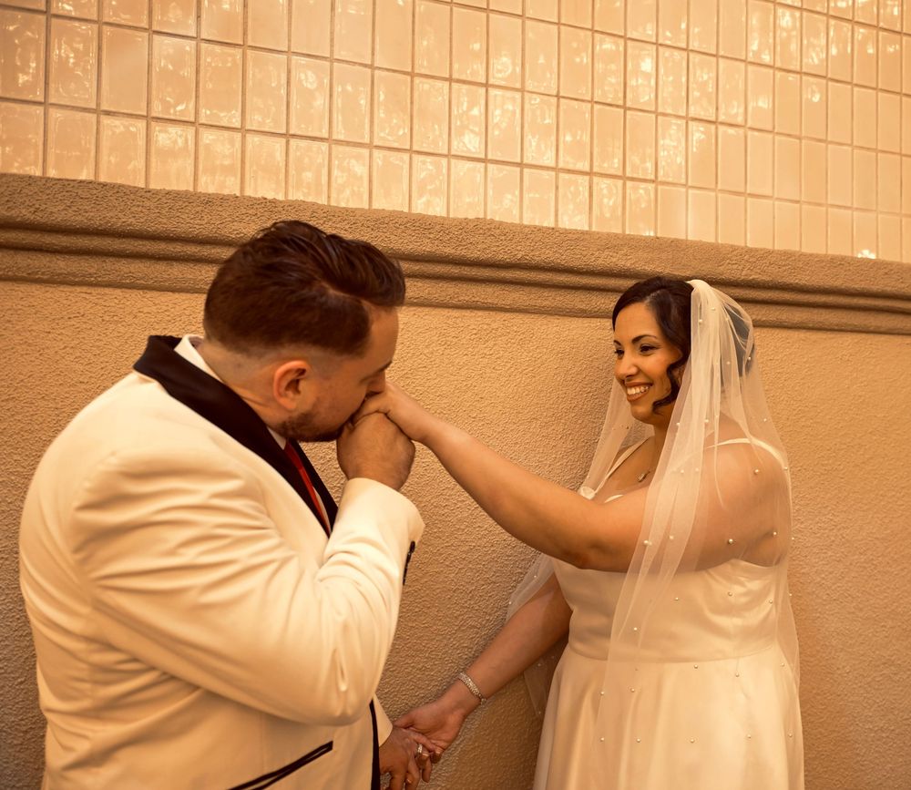 Groom in a white tuxedo jacket kisses his bride's hand as she smiles in her veil against a warm tiled wall.