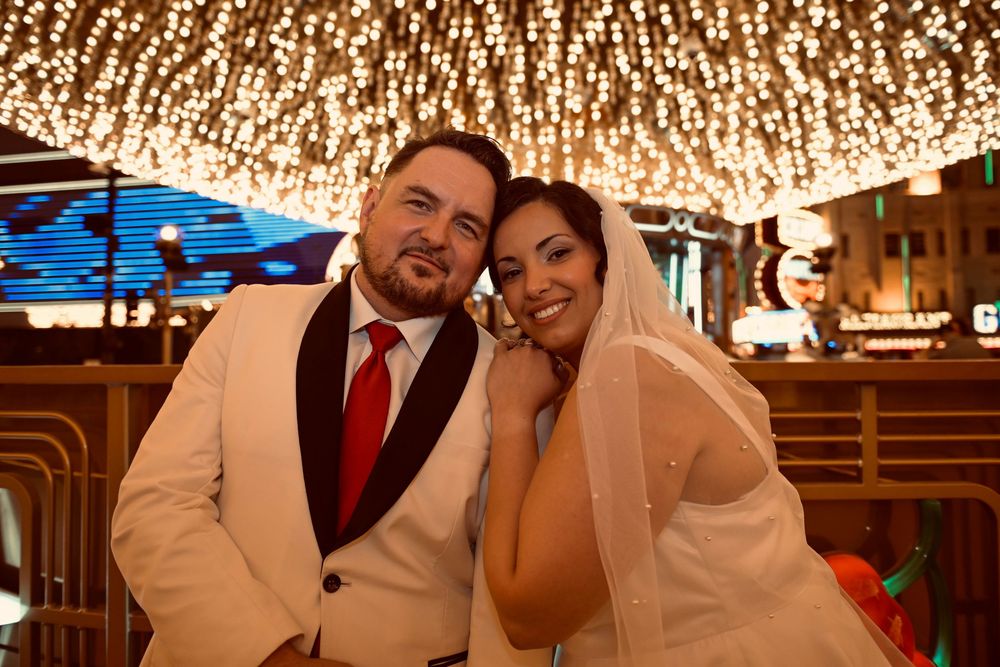 Smiling bride in a white veil leans her head against a groom in a white tuxedo jacket and red tie under glowing string lights at night.