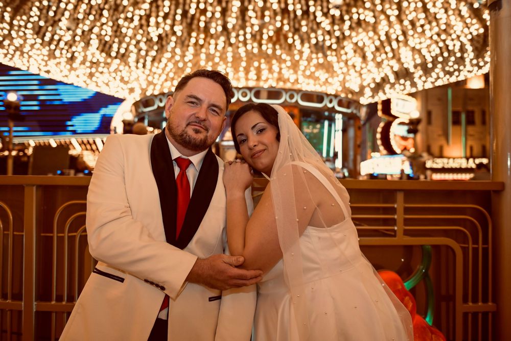 Smiling groom in a white tuxedo jacket and red tie embraces his bride in a veil under a sparkling canopy of lights at a Las Vegas wedding.