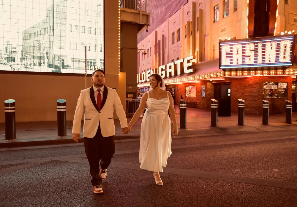 Newlywed couple holding hands in wedding attire as they walk down a Las Vegas street at night past glowing casino neon signs.