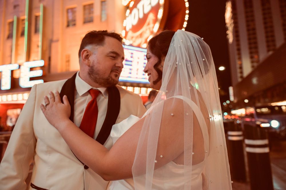 Bride in a white dress and long veil embraces her bearded groom in a cream suit and red tie beneath glowing neon signs at night.