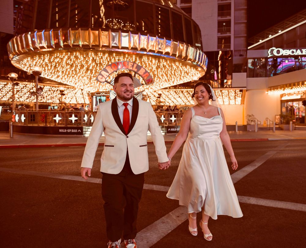Bride in a white dress and groom in a cream tuxedo jacket holding hands on a street in front of glittering Las Vegas casino lights at night.