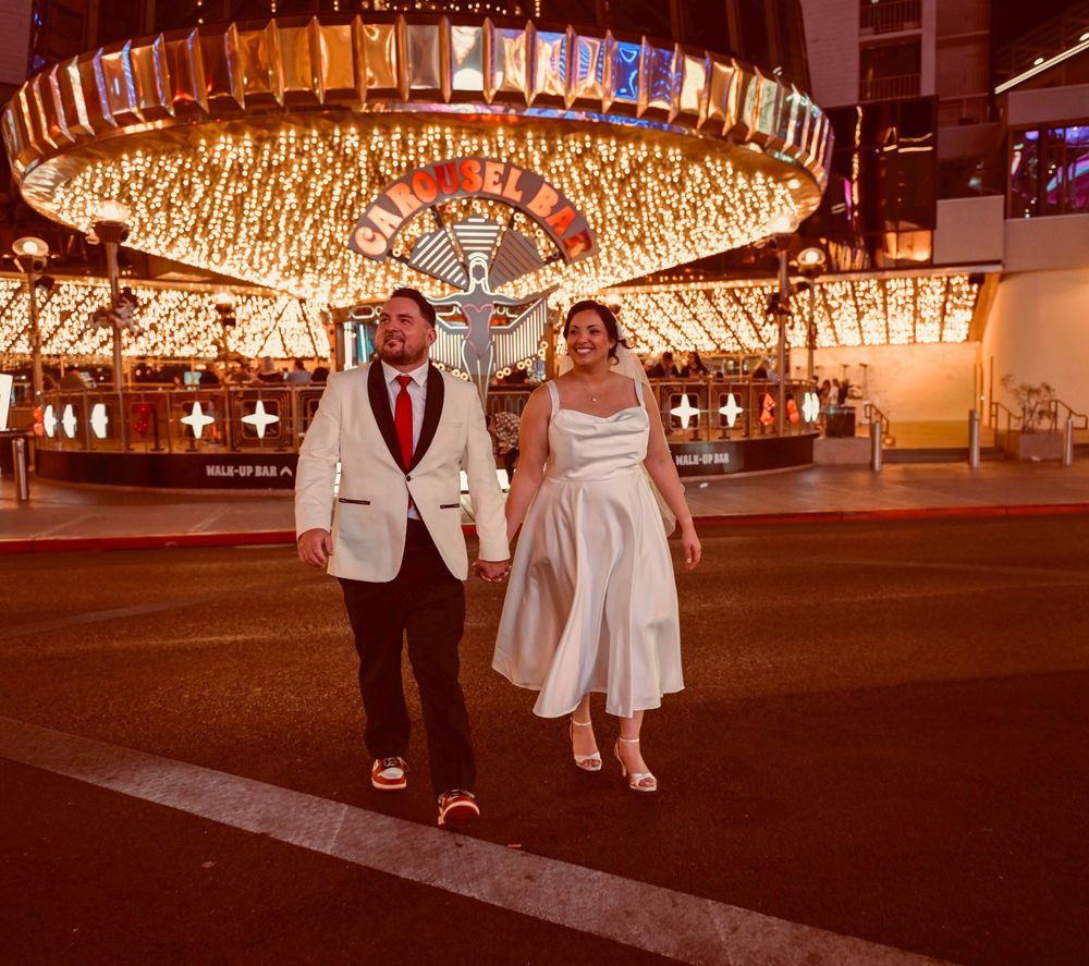 Bride in a short white dress and groom in a white jacket with red tie walk hand in hand past glittering casino lights in Las Vegas at night.