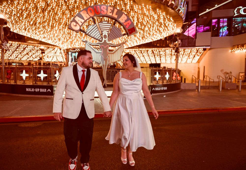 Couple in a white blazer and white dress holding hands under the glowing Fremont Street canopy lights in downtown Las Vegas at night.