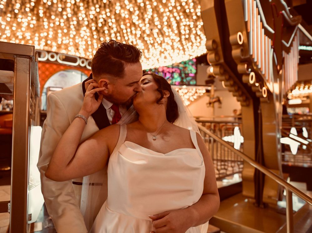 Bride in a white gown and groom in a cream jacket share a kiss beneath glowing string lights at an indoor wedding venue.