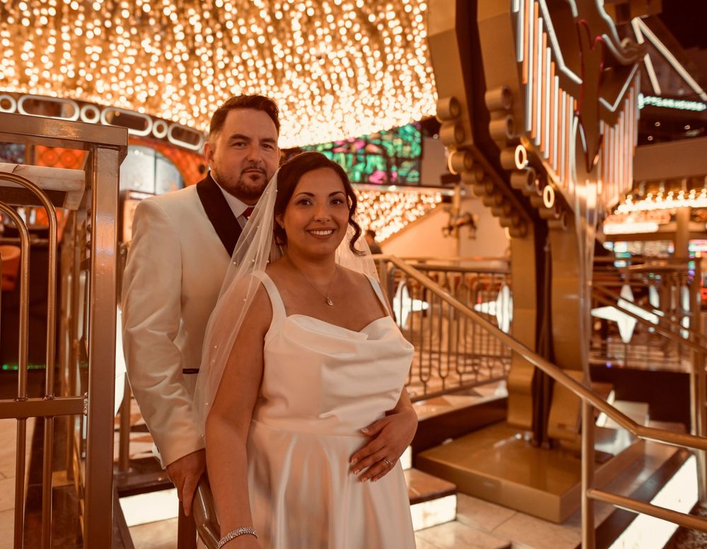 Smiling bride in a white dress and veil poses with her groom in a white jacket beside a glittering staircase in a Las Vegas casino.