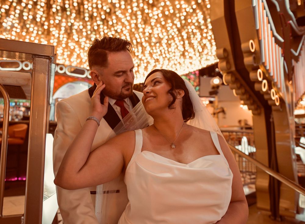 Groom in a cream suit embraces his smiling bride in a white gown and veil beneath a ceiling of sparkling golden lights.