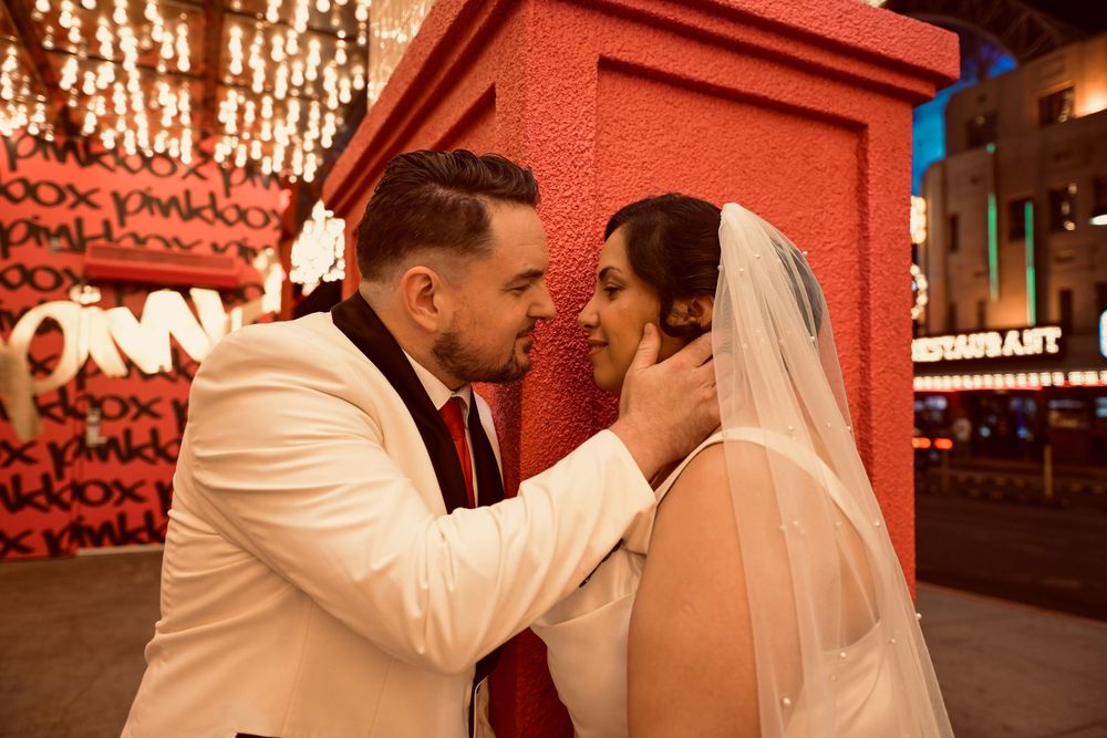 Bride in a veil and groom in a white tuxedo jacket share an intimate moment beside a red pillar with glowing city lights at night.