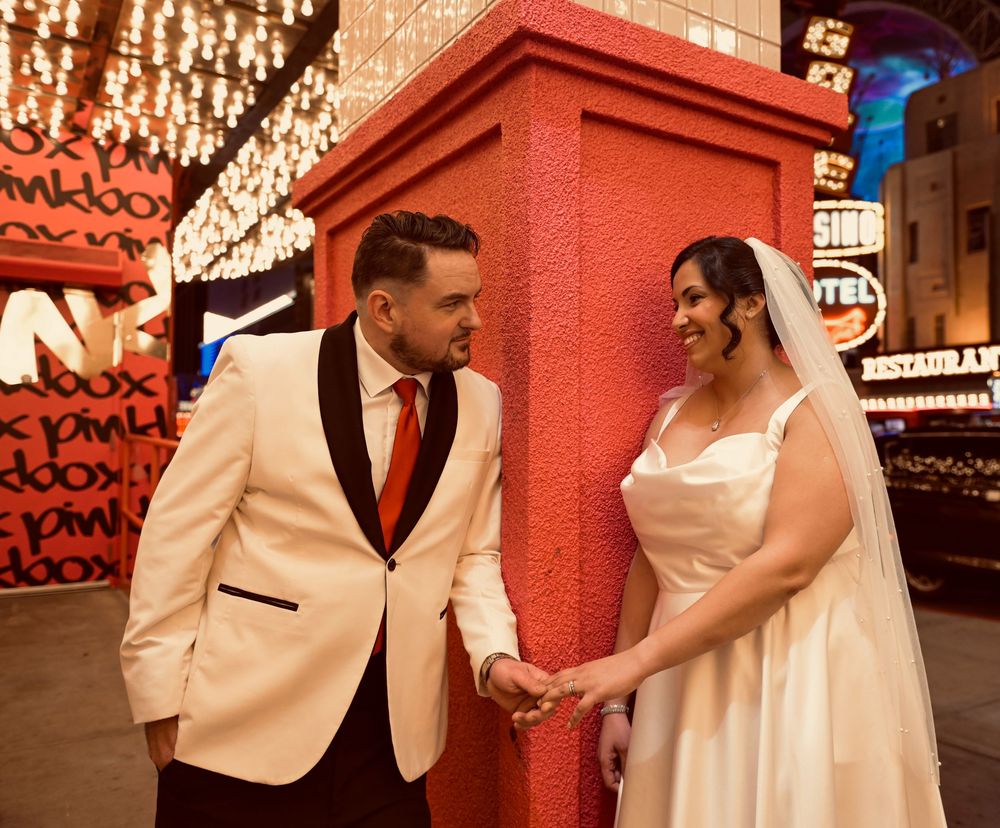 Bride in a white gown and veil holds hands with a groom in a cream tuxedo jacket and red tie outside glowing Las Vegas casino signs.