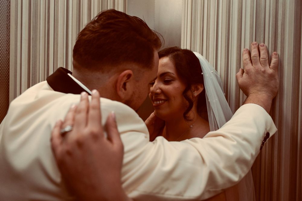 A smiling bride in a white veil shares an intimate moment with her groom as they embrace against a softly lit, curtained wall.