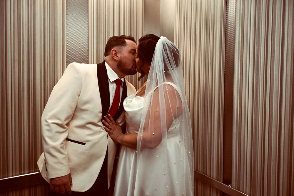 Bride in a white gown and veil shares a wedding kiss with her groom in a cream tuxedo jacket inside an elegant striped elevator.